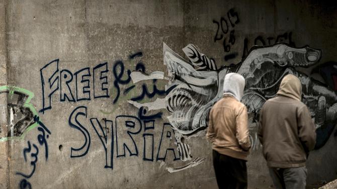 Migrants walk past graffiti reading &quot;Free Syria&quot; in the &quot;Jungle&quot; migrant camp in Calais, northern France, on February 12, 2016