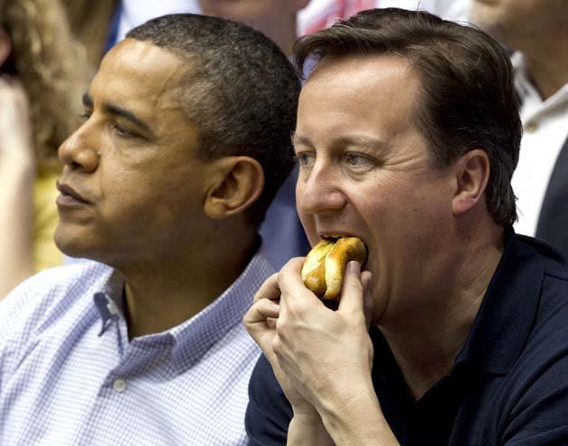 رئيس الوزراء البريطاني ديفيد كاميرون يحشر سندويتش من الهوت دوج في فمه خلال مشاهدة مبارة رياضية برفقة أوباما