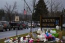 Cars drive below a U.S. flag at half mast and past a memorial outside the entrance to Newtown High School in Newtown, Connecticut